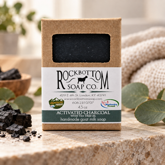 Rock Bottom Soap Co. activated charcoal goat milk soap with tea tree oil in kraft packaging, displayed with eucalyptus and charcoal on a natural stone surface.